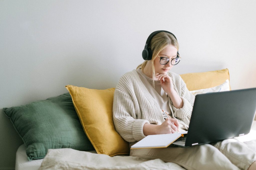 Junge Frau mit blonden Haaren und Kopfhörern sitzt auf dem Sofa und arbeitet am Laptop