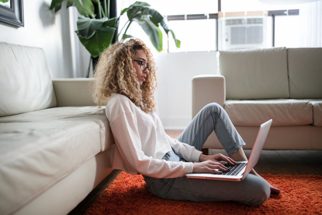 Frau mit blonden lockigen Haaren und Brille sitzt vor dem Sofa und schaut auf einen Laptop
