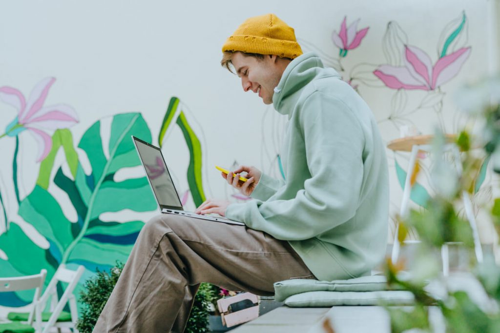 Junger Mann mit bunten Klamotten arbeitet draußen mit Handy und Laptop