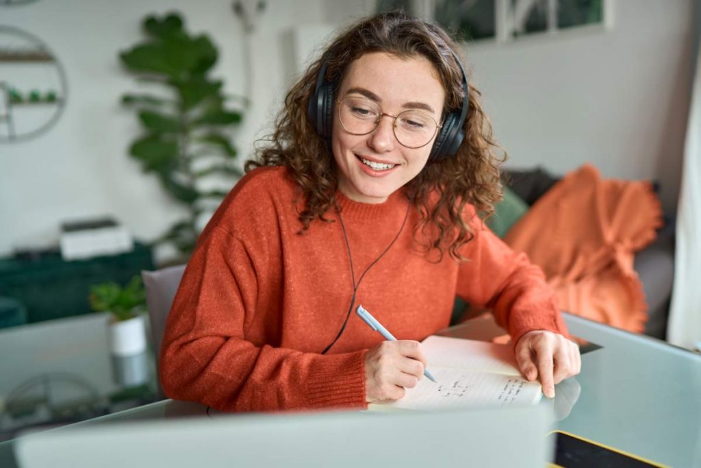 Eine junge, lächelnde Frau im orangefarbenem Pullover schreibt in ihr Notizheft