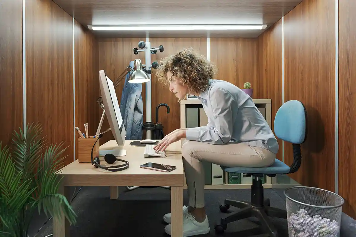 Frau mit lockigen Haaren sitzt mit krummen Rücken an einem zu kleinen Schreibtisch in einem kleinen Büro