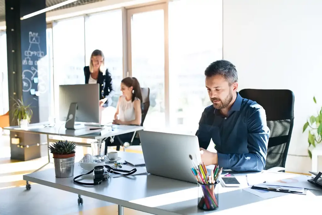 Mitarbeitende sitzen in einem offenen, hellen Büro und arbeiten an ihren Schreibtischen. Frau im Hintergrund steht